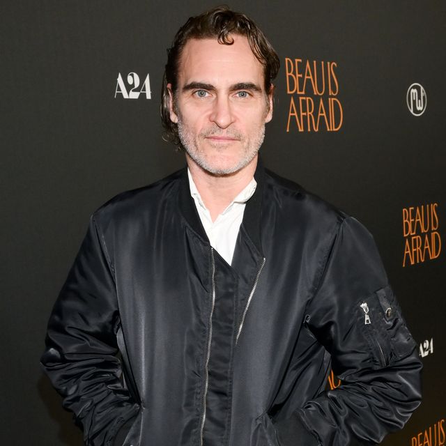 joaquin phoenix smiles at the camera, he wears a black jacket and white collared shirt, his hands are in his jacket pockets and he stands in front of a black background