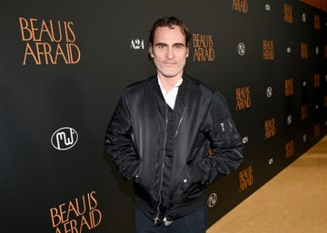 joaquin phoenix smiles at the camera, he wears a black jacket and white collared shirt, his hands are in his jacket pockets and he stands in front of a black background