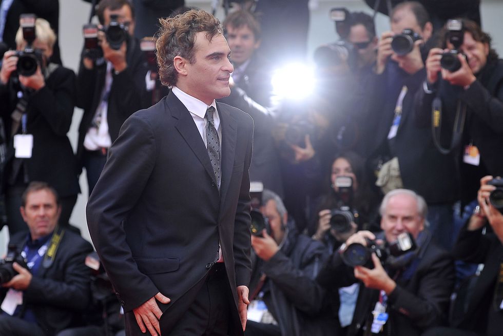 joaquin phoenix walks past a slew of photographers and smiles, he wears a black suit with a white collared shirt and gray and black patterned tie
