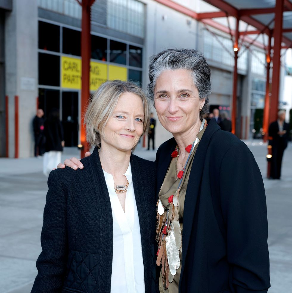 jodie foster and alexandra hedison stand next to each other, smiling and hugging for a photo, they bothe wear dark jackets over blouses