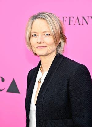 jodie foster looks at the camera with a small smile, she wears a black blazer, white shirt, and gold necklace, she stands in front of a hot pink backdrop