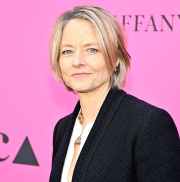 jodie foster looks at the camera with a small smile, she wears a black blazer, white shirt, and gold necklace, she stands in front of a hot pink backdrop