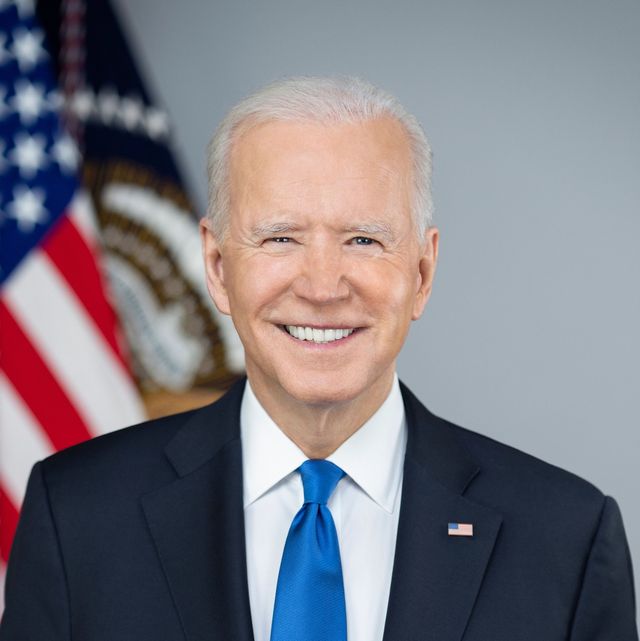 joe biden smiles at the camera while standing in front of a blurry american flag, he is wearing a navy suit, a white collared shirt, a royal blue tie, and an american flag pin on his lapel, he has a white pocket square in his breast pocket