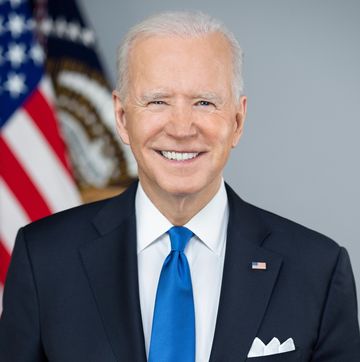 joe biden smiles at the camera while standing in front of a blurry american flag, he is wearing a navy suit, a white collared shirt, a royal blue tie, and an american flag pin on his lapel, he has a white pocket square in his breast pocket