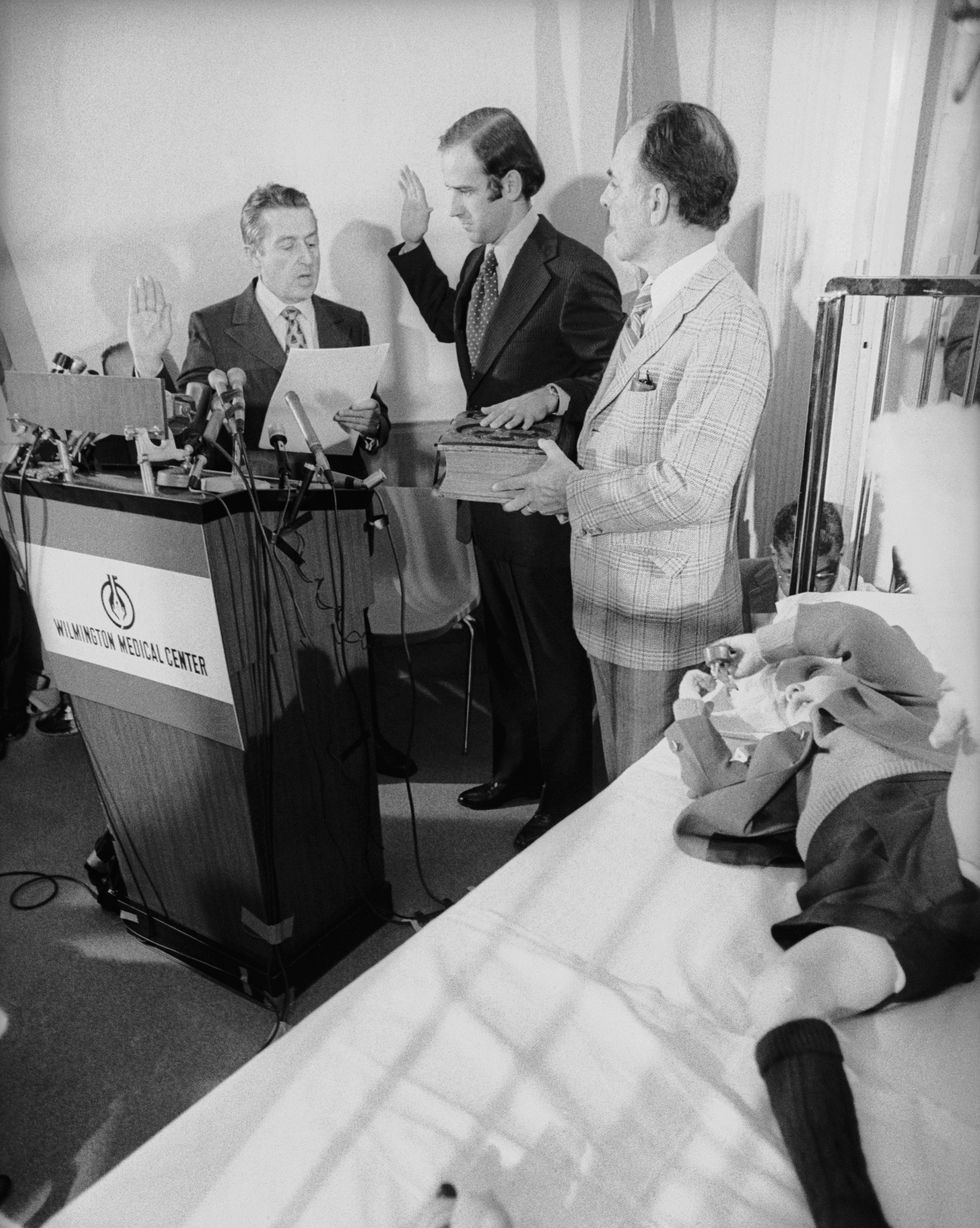 joe biden stands between two men who help swear him in behind a wooden podium, young beau biden rests on a hospital bed nearby