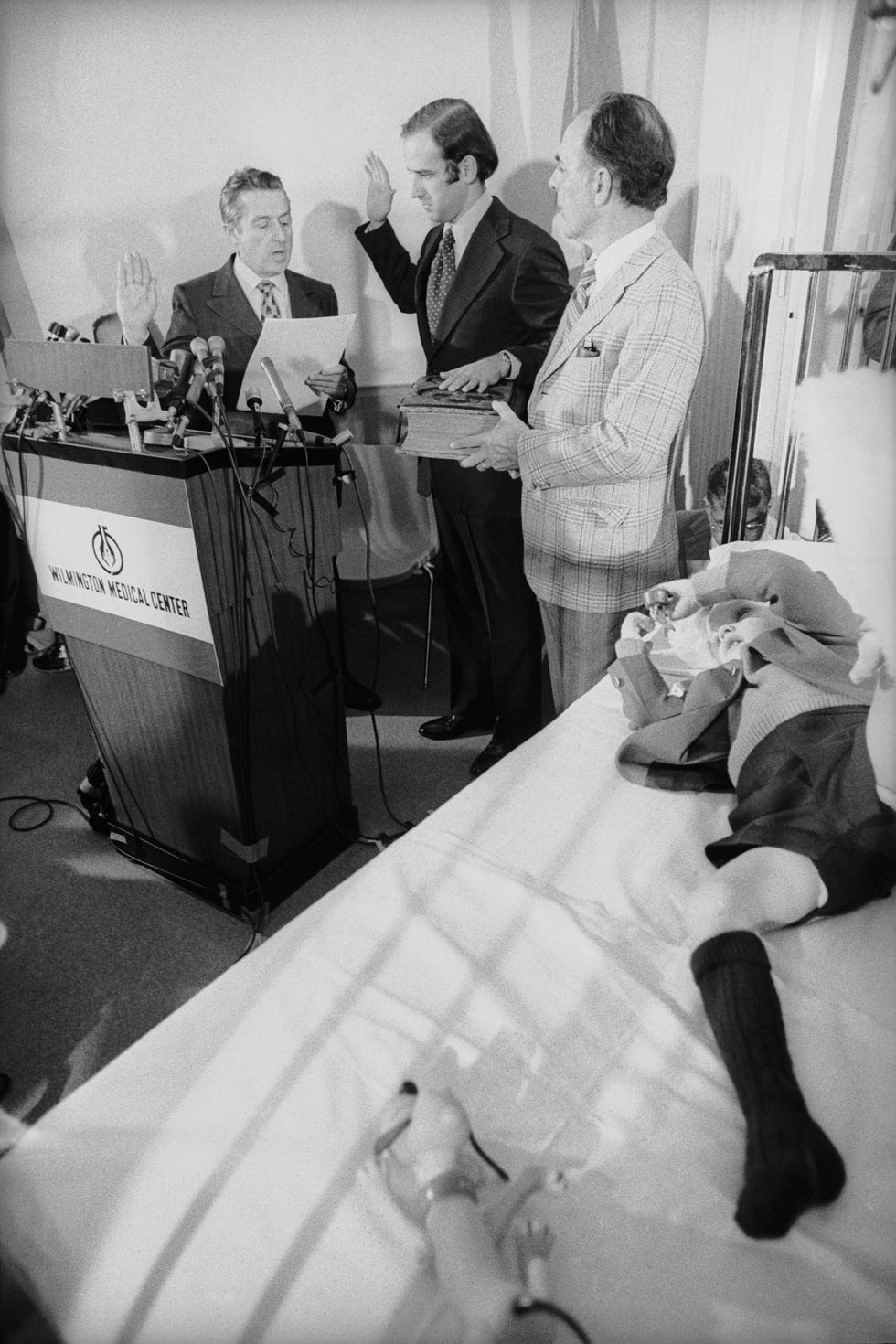 joe biden stands between two men who help swear him in behind a wooden podium, young beau biden rests on a hospital bed nearby