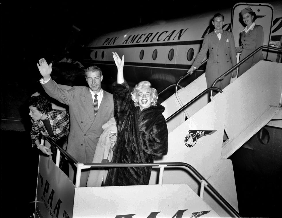 joe dimaggio and marilyn monroe stand on a staircase next to a plane, they wave and smile