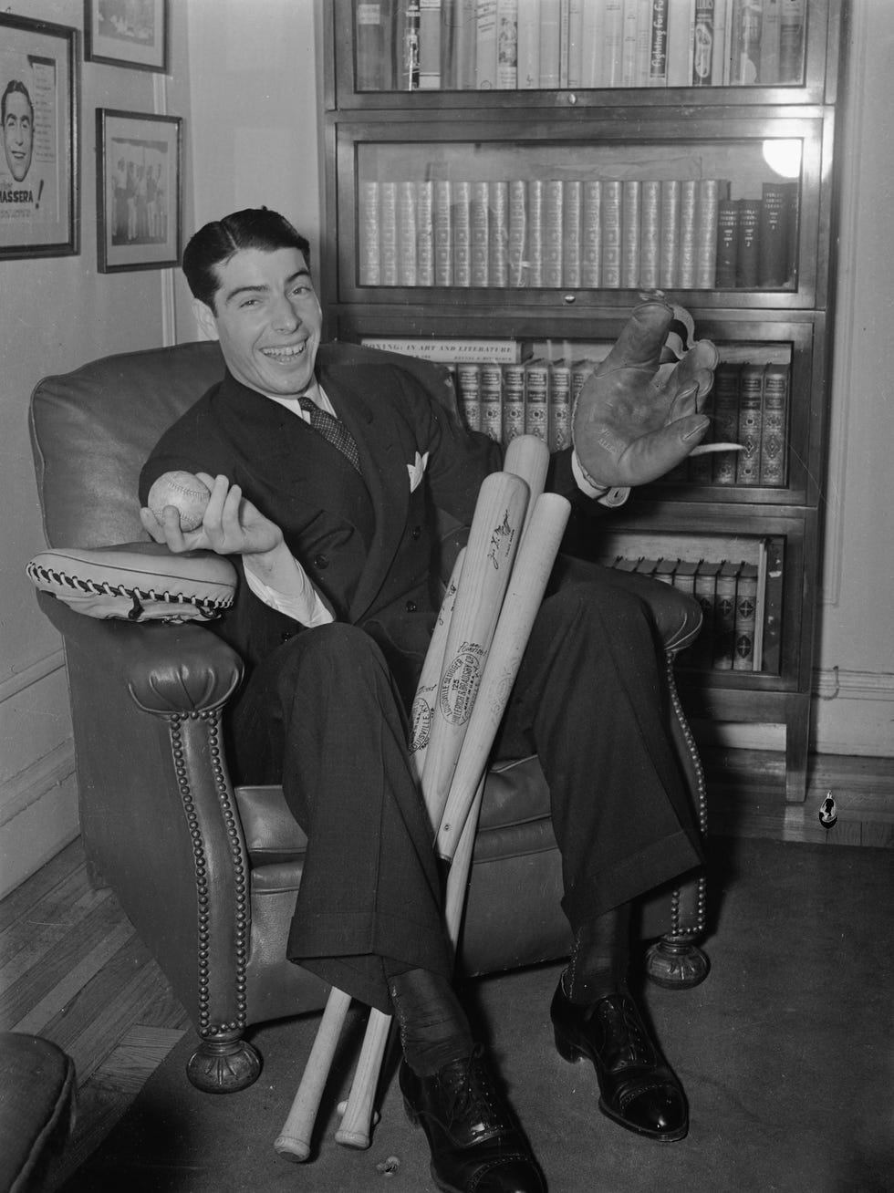 joe dimaggio sits in an armchair wearing a glove and holding a ball, several bats rest between his legs
