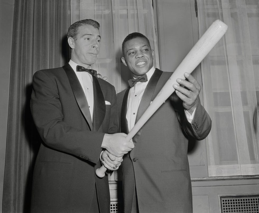 joe dimaggio and joe dimaggio look at a bat that dimaggio holds, both men wear suits