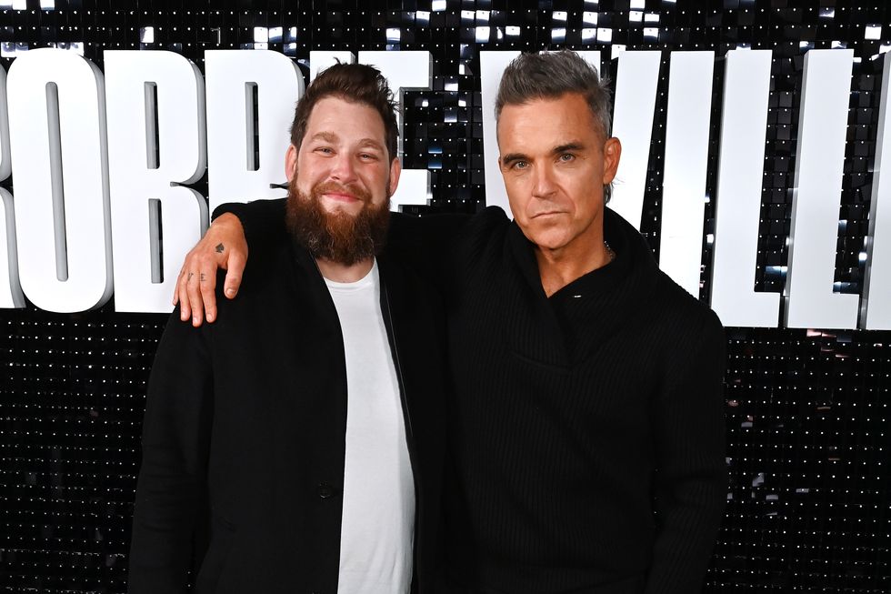 Joe Pearlman y Robbie Williams posan abrazados frente a letras gigantes que deletrean Robbie Williams.