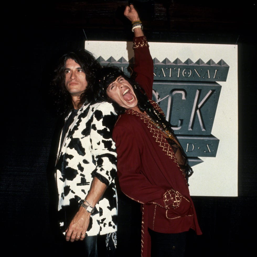 joe perry and steven tyler stand back to back in front of a poster, perry is wearing a cow print suit jacket and black pants, tyler is wearing a red jacket with gold accents and has one arm thrust over his head in the arm