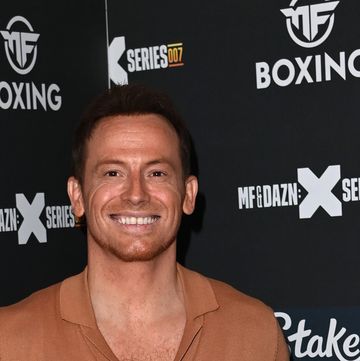 joe swash, a young man stands smiling at the camera and standing with hands behind his back, he wears black jeans with a brown tshirt