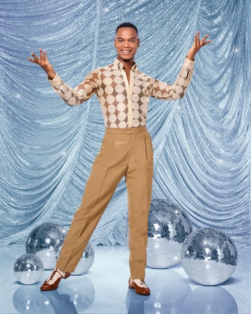 johannes radebe smiles and holds up his arms as he stands in front of a glittery blue fabric backdrop, with disco balls on the floor behind him