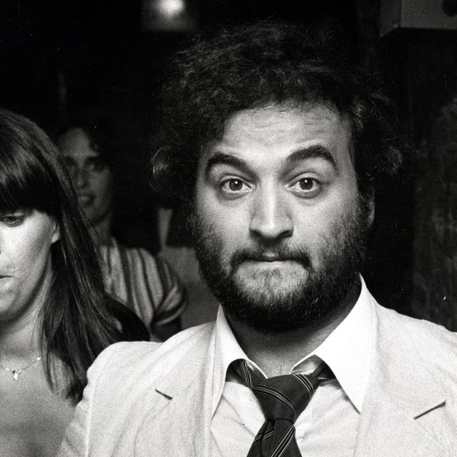 john belushi looks at the camera, he wears a light colored suit with a striped tie