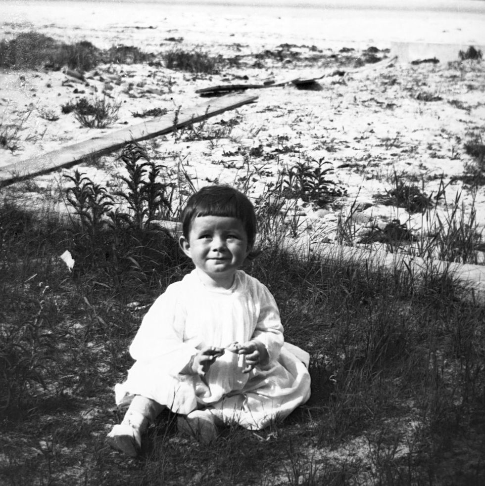 infant john f kennedy sits on grass and smiles, behind him is a body of water