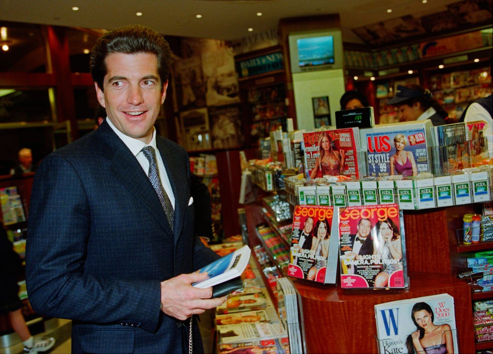 john f kennedy jr stands next to a magazine display and holds a book and a wallet, he looks to the left and wears a navy suit jacket, white dress shirt, and dark colored tie