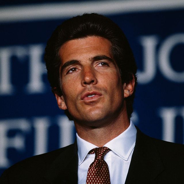 john f kennedy jr stands and speaks, he wears a black suit jacket, white dress shirt and red polka dot tie