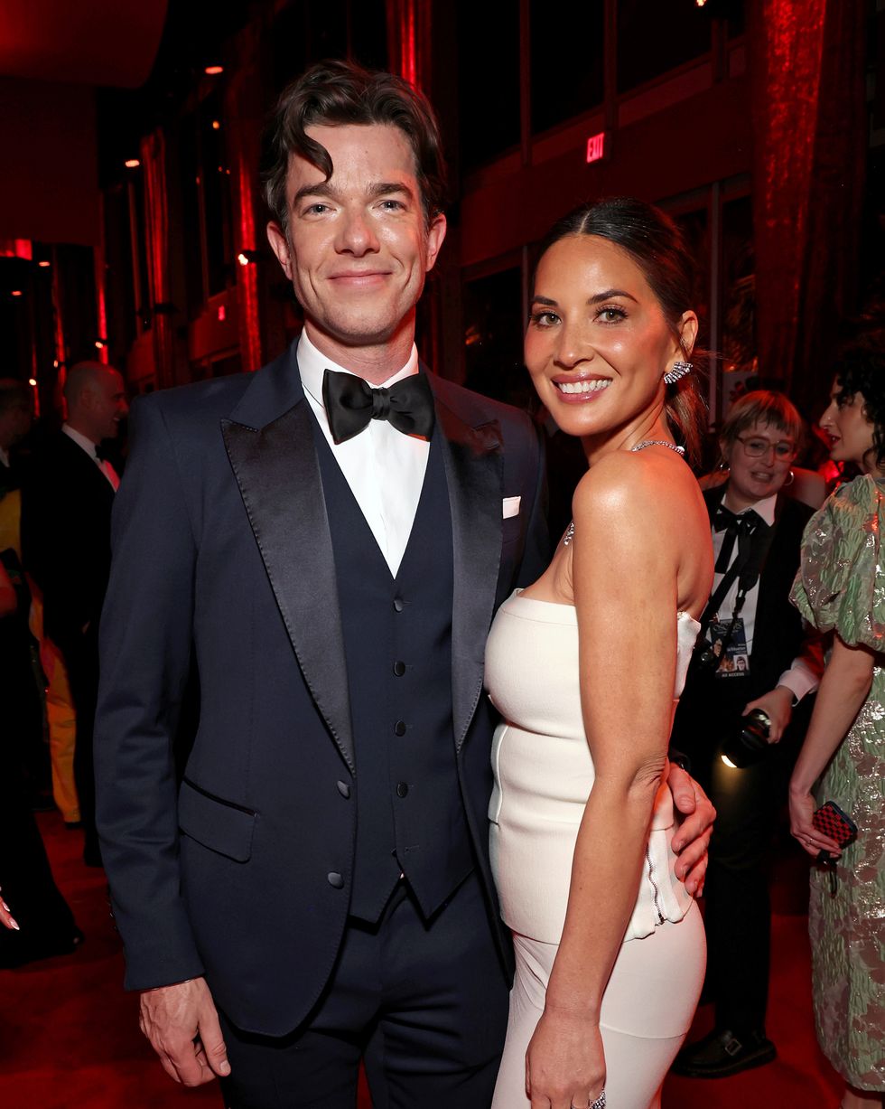 john mulaney and olivia munn pose for a photo in a crowded room, he holds a hand on her waist and wears a navy tuxedo, she wears a white strapless gown