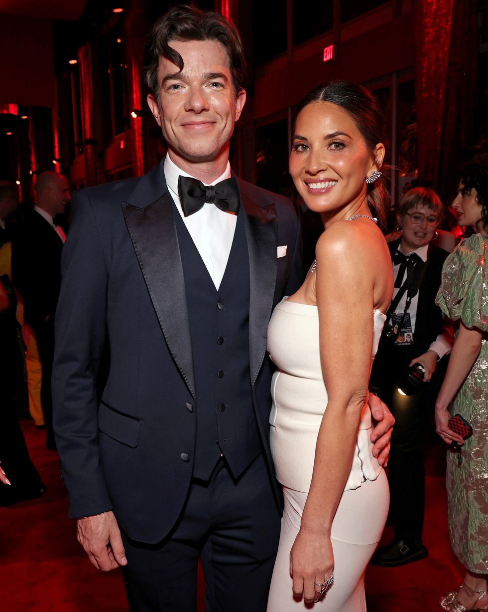 john mulaney and olivia munn pose for a photo in a crowded room, he holds a hand on her waist and wears a navy tuxedo, she wears a white strapless gown