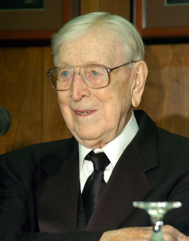 john wooden wears a black suit jacket, black tie, white collared shirt, and aviator style eye glasses, he looks off camera to the left and is smiling slightly
