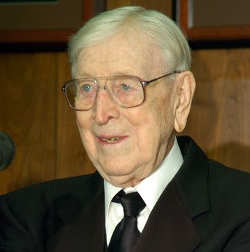john wooden wears a black suit jacket, black tie, white collared shirt, and aviator style eye glasses, he looks off camera to the left and is smiling slightly