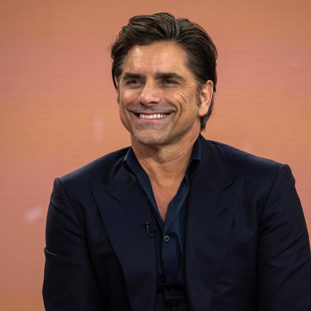 john stamos looks left of the camera and smiles, he wears a navy suit jacket and dark blue collared shirt