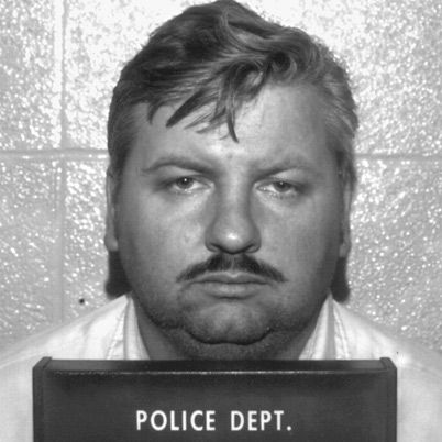 a mugshot of john wayne gacy who looks directly at the camera with a neutral expression on his face, below his chin is a black marker that reads "police dept"