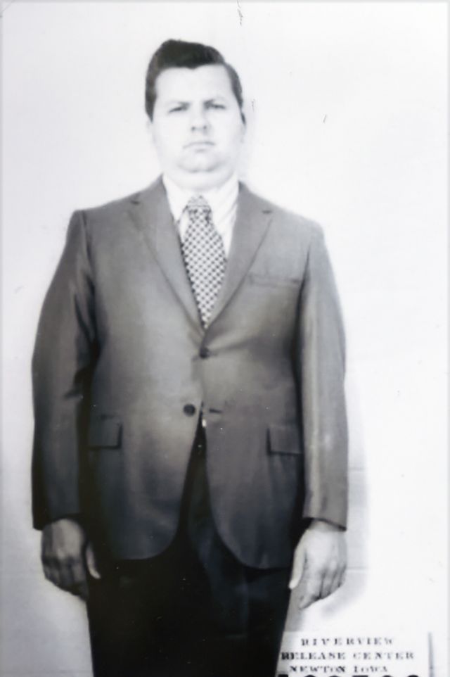 a black and white photo of john wayne gacy, who wears a suit and stands straight, looking ahead