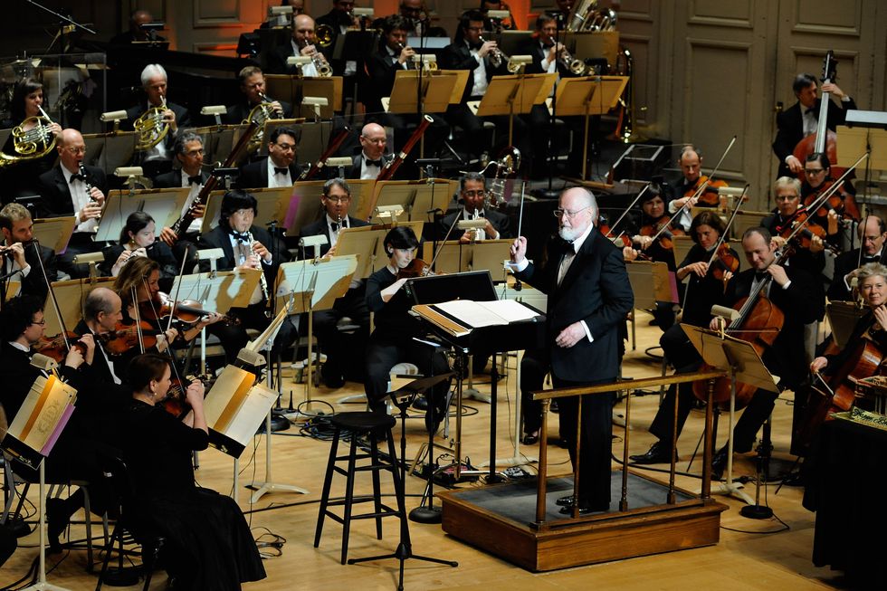 john williams stands on a platform and waves a baton while orchestra musicians play around him
