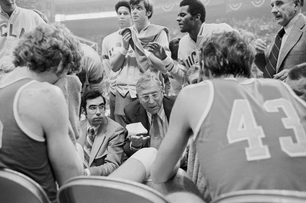 a black and white photo of several basketball players and assistant coaches surrounding john wooden, who talks to them
