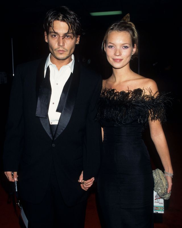 johnny depp and kate moss stand and hold hands while looking at the camera, he wears a black tuxedo with an untied bowtie, she wears a black strapless dress with feather fringe at the neckline