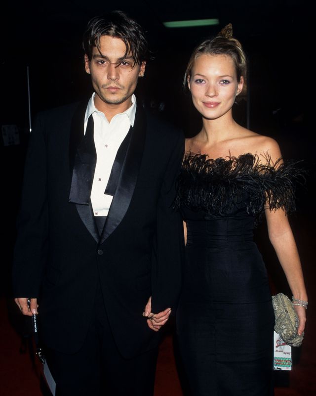 johnny depp and kate moss stand and hold hands while looking at the camera, he wears a black tuxedo with an untied bowtie, she wears a black strapless dress with feather fringe at the neckline