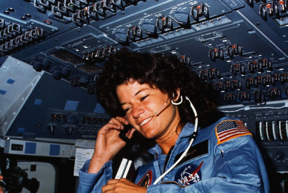 a woman smiles as she holds a tape recorder wears a headset and looks down, she is inside a small enclosed area with many buttons and nobs