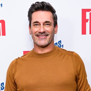 jon hamm, a man stands smiling at the camera, brown hair and light facial hair, wearing an orange jumper and blue jeans