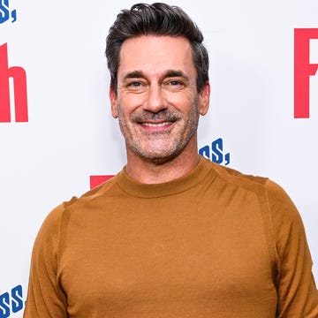 jon hamm, a man stands smiling at the camera, brown hair and light facial hair, wearing an orange jumper and blue jeans