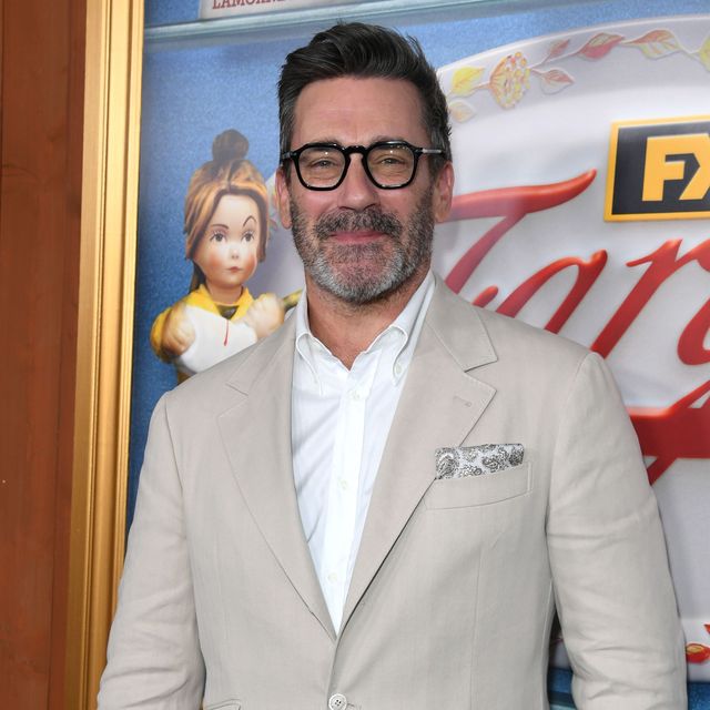 jon hamm smiles at the camera while standing in front of a fargo tv show background, he wears a tan suit with a white collared shirt and patterned pocket square