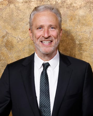 jon stewart smiles at the camera, he wears a black suit jacket, white collared shirt, and a dark striped tie