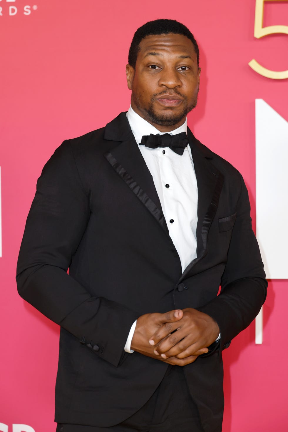 Jonathan Majors, un hombre de pie mirando a la cámara con las manos juntas, cabello negro muy corto, vestido con un traje negro y pajarita con camisa blanca.