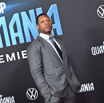 jonathan majors stands in a gray suit, white shirt, and dark gray tie, behind him is a black background for ant man and the wasp quantumania