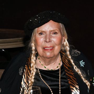 joni mitchell smiles at the camera, she wears a black beaded hat, black top and black sweater with tiger print trim and embroidered plant leaves