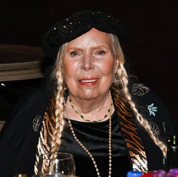 joni mitchell smiles at the camera, she wears a black beaded hat, black top and black sweater with tiger print trim and embroidered plant leaves