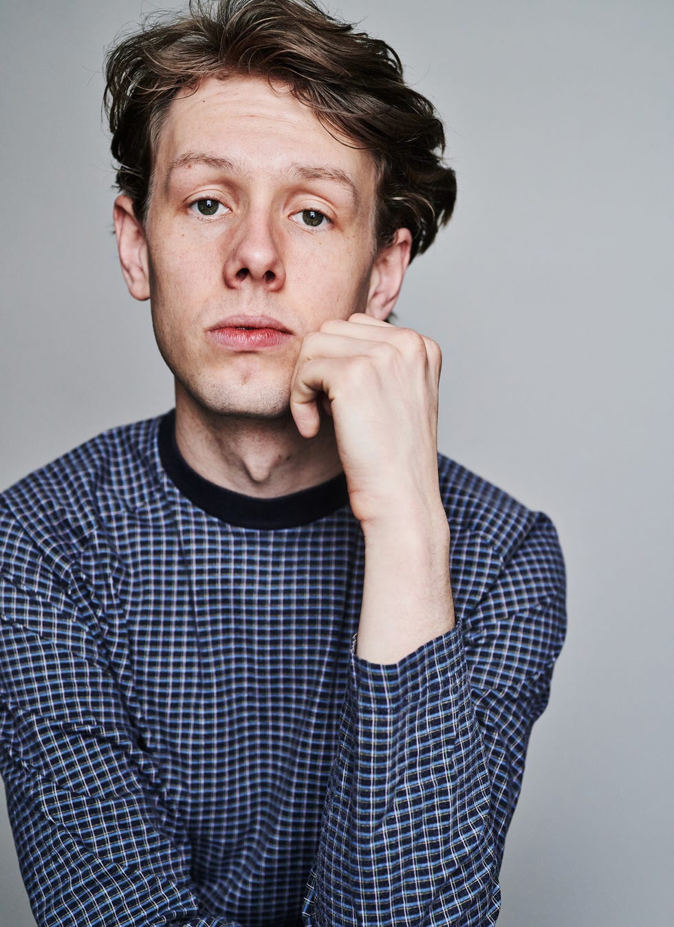Portrait of a person with one hand raised to their chin wearing a patterned longsleeve shirt