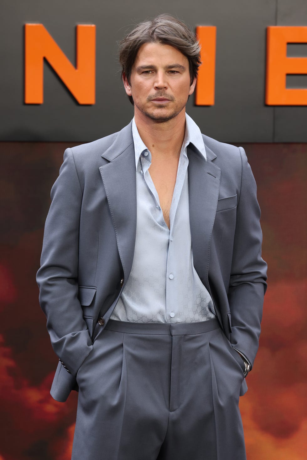 josh hartnett stands with his hands in his pockets, he wears a blue gray suit with a light blue shirt unbuttoned at the collar