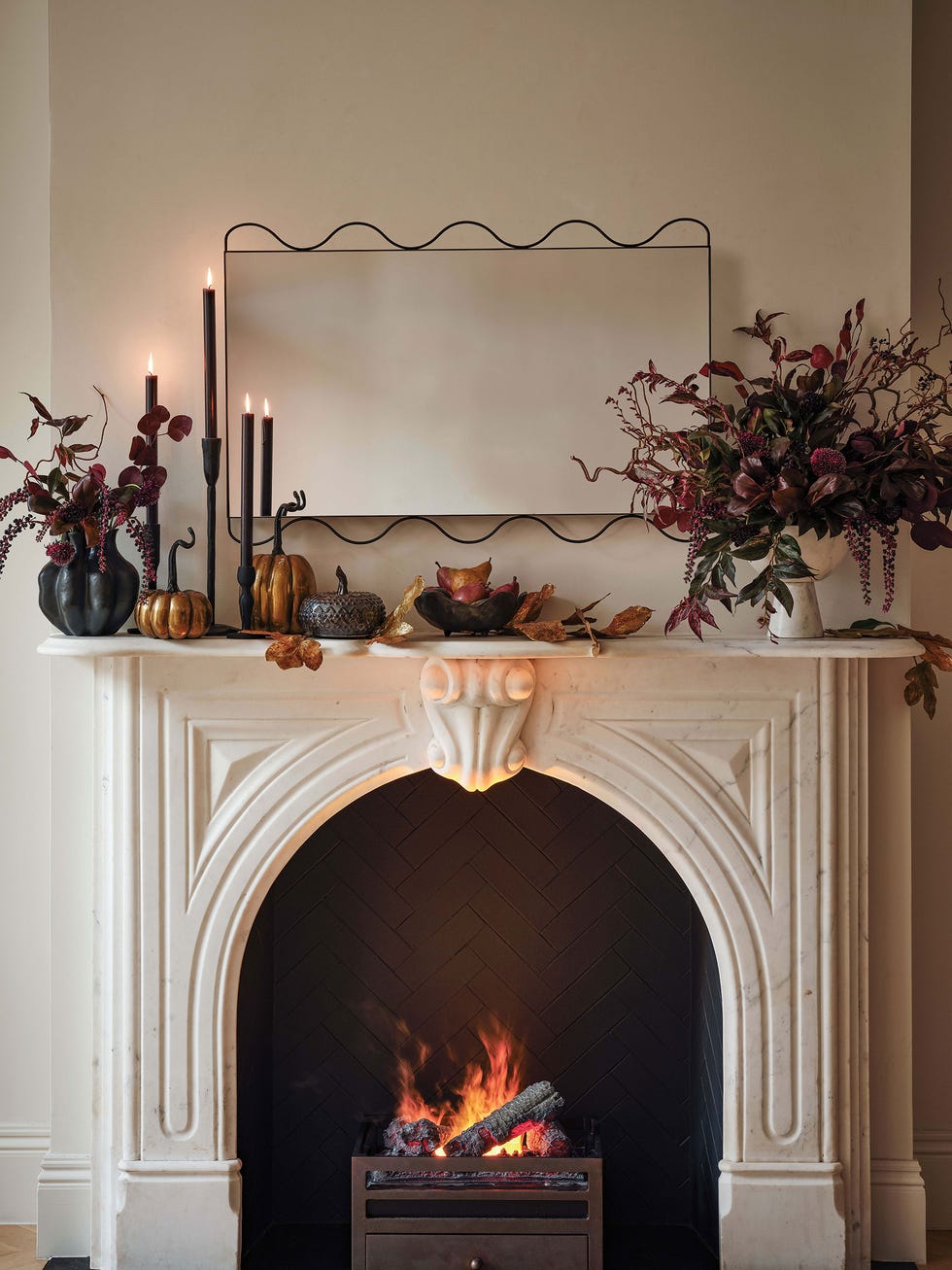 a marble fireplace features a classic design with an arched opening containing a glowing fire above the mantle is a wavy edged black mirror the mantle is decorated with tall, slender black candles, various decorative gourds, a vase with dark foliage, and dried leaves, creating an autumnal atmosphere