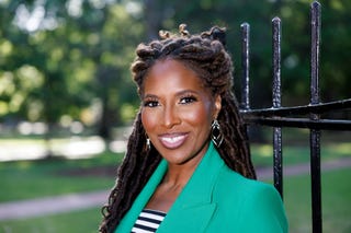 person with dreadlocks wearing a green blazer standing next to a black fence in a park
