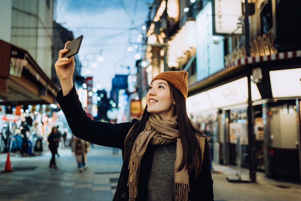 joyful young asian female traveller taking a selfie with smartphone while exploring and strolling in busy downtown city street at night in osaka, japan lifestyle and technology travel, vacation and holiday concept
