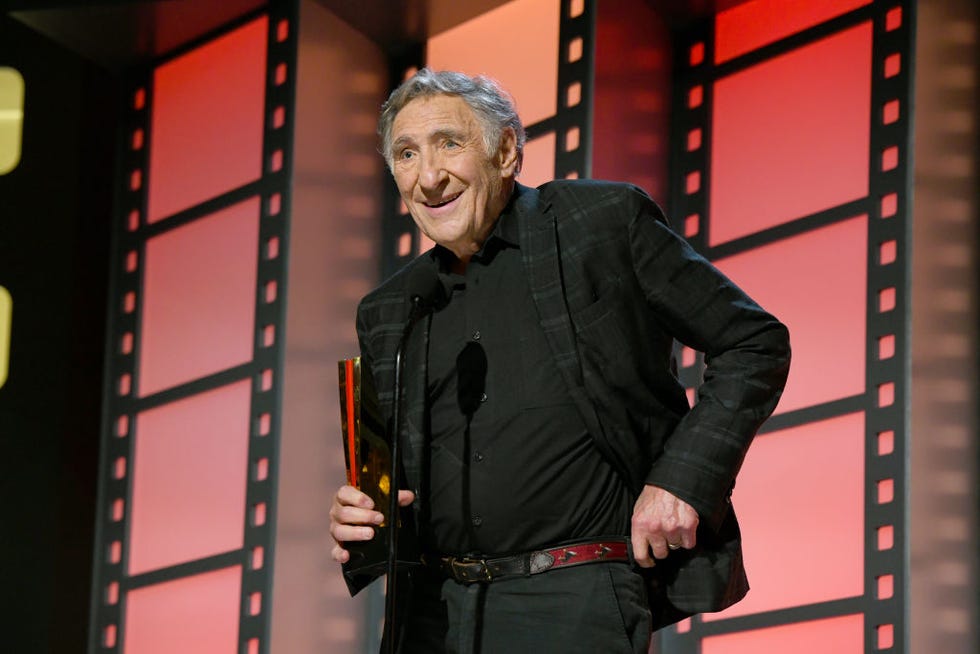 judd hirsch, wearing a black suit jacket and shit, smiling and speaking into a microphone on a stage while holding an award