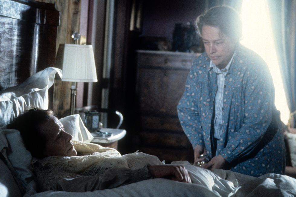 a woman lies in bed as another woman stands on the side of the bed and looks at her, sun streams into the room through a partially open curtain behind
