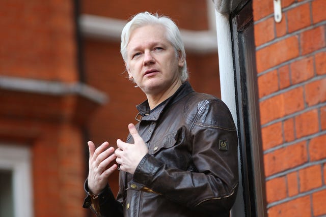 julian assange standing in front of a brick building and gesturing with his hands as he speaks