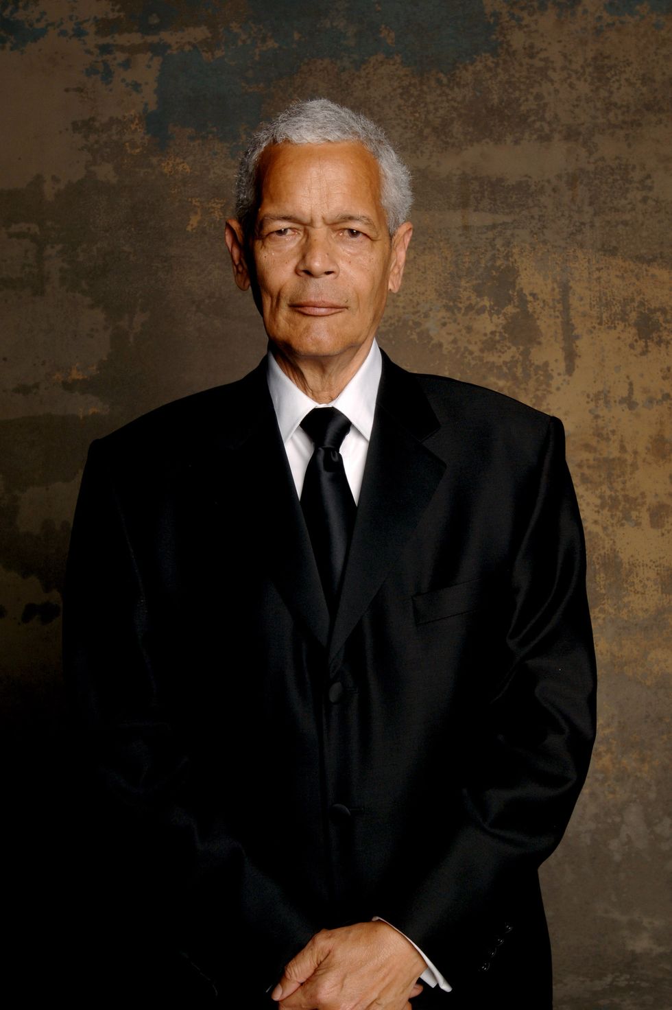 julian bond stands and looks at the camera with his hands clasped in front of him, he wears a black suit and tie with a white collared shirt
